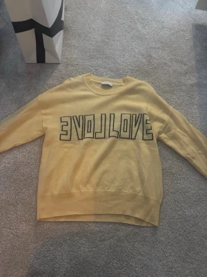 Gul stickad tröja från Sandro Paris - Trendig gul stickad tröja från Sandro Paris med svart texten 'EVOL/LOVE' framtill. Rund halsringning och ribbade muddar vid ärmslut och nederkant. Perfekt för dig som vill ha en cool statementtröja i garderoben. 100% cashmere