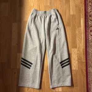 Gråa mjukisbyxor från Adidas - Säljer ett par gråa mjukisbyxor från Adidas med svarta ränder på benen och Adidas-logga på ena låret. Byxorna har resår i midjan och raka ben för en avslappnad look. 