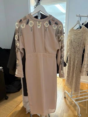 Beige klänning med paljettdetaljer - Säljer en beige långklänning med långa ärmar och dekorativa paljetter och pärlor på axlar och ärmar. Klänningen har en rak passform och rund halsringning. Perfekt för dig som vill sticka ut med eleganta detaljer.