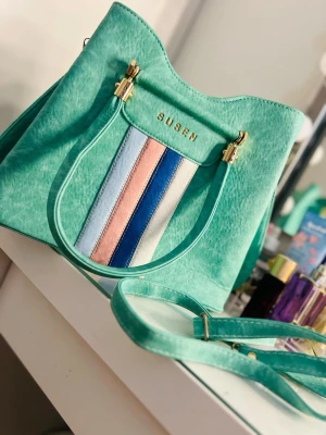 Mintgrön handväska från Susen - Trendig mintgrön handväska från Susen med guldfärgade detaljer och dekorativa ränder i ljusblått, rosa, mörkblått och vitt på framsidan. Väskan har både handtag och axelrem, perfekt för dig som gillar färg och stilren design. Materialet är konstläder.