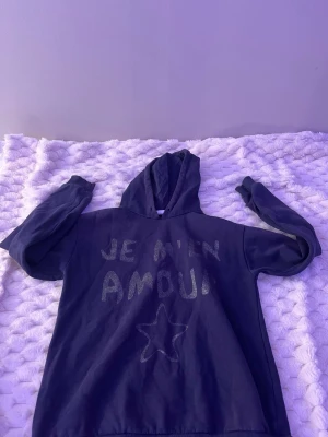 Mörkblå hoodie med text och stjärna - Cool mörkblå hoodie med huva och trycket 'JE M'EN AMOUR' i glittriga bokstäver på bröstet samt en glittrig stjärna nedtill. Hoodien har långa ärmar och en avslappnad passform. Perfekt för dig som gillar statement-plagg och vill sticka ut lite extra.