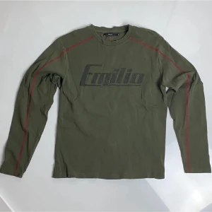 Olivgrön långärmad tröja Emilio - Olivgrön långärmad tröja från Emilio med tryckt logga i svart framtill. Tröjan har röda sömdetaljer längs axlar och ärmar samt rund halsringning. Tillverkad i mjukt bomullsmaterial för en avslappnad look.