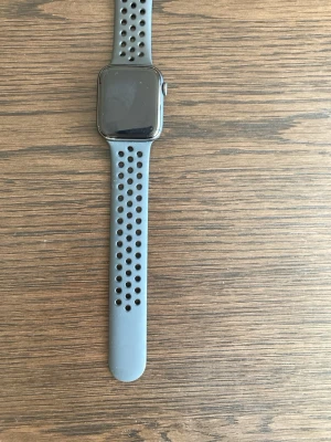 Apple Watch series 6 med mörkgrå/svart sportarmband - Apple Watch med svart fyrkantig urtavla och mörkgrå/svart silikonarmband (ser ljusgrå ut på bilden) med svarta hål. Armbandet är sportigt och bekvämt, perfekt för träning och vardag. Klockan har en modern och stilren design som passar till det mesta.