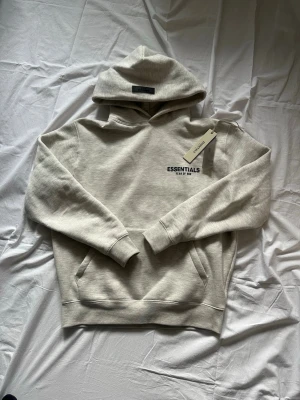Beige Essentials hoodie med huva - Säljer en beige hoodie från Essentials Fear of God med huva och känguruficka. Diskret logga på bröstet och mjukt material som känns riktigt skönt. Perfekt oversized passform och stilren look. Levereras i originalpåse med tag kvar.