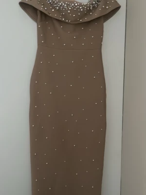 Beige långklänning med pärlor - Elegant beige långklänning med offshoulder-design och dekorerad med pärlor och strass över hela klänningen. Klänningen har en smickrande, figurnära passform och är perfekt för speciella tillfällen. Materialet känns mjukt och följsamt.