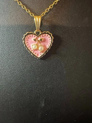 Halsband med hjärthänge och pärlor - Gulligt halsband med guldfärgad kedja och ett hjärthänge i rosa emalj. Hänget har en körsbärsdetalj med två små pärlor och gröna blad. Perfekt accessoar för dig som gillar söta och lekfulla smycken.