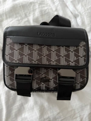 Lacoste väska  - Snygg handväska från Lacoste i brunt och svart med grafiskt mönster och vita detaljer. Väskan har ett lock med två spännen, justerbar axelrem och präglad Lacoste-logga på framsidan. Materialet är konstläder och textil, perfekt för dig som gillar streetstyle.