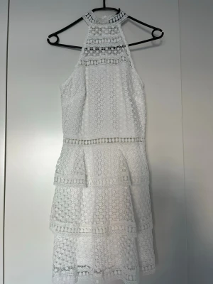 Vit spetsklänning med hög hals - Vit ärmlös kortklänning med hög hals och flera lager volanger. Klänningen har vackra spetsdetaljer och ett elegant, strukturerat mönster. Perfekt för sommarens alla tillfällen och ger en romantisk vibe.