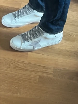Vita Golden Goose sneakers - Säljer ett par vita Golden Goose sneakers i läder med grå skosnören och den ikoniska stjärnan på sidan. Skorna har en låg silhuett och detaljer i mocka på hälen. Perfekta för dig som gillar en clean men ändå unik look. (Rep)