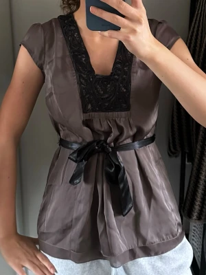 Brun blus med spetsdetalj och sidenband - Snygg brun blus med svart spetsdetalj vid ringningen och ett matchande svart sidenband i midjan. Blusen har en lätt glansig finish och korta ärmar. Perfekt för dig som gillar feminina detaljer och en markerad midja.
