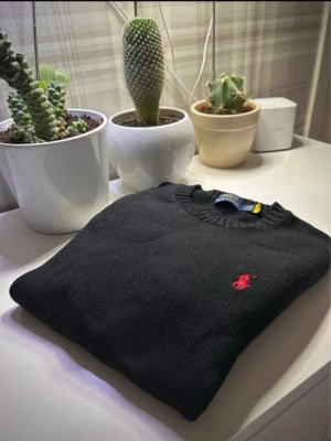 Svart stickad tröja från Polo Ralph Lauren - Svart stickad tröja från Polo Ralph Lauren med klassisk rund halsringning och röd broderad logotyp på bröstet. Tröjan är tillverkad i ett mjukt stickat material och har ribbade muddar vid halsen. Perfekt för dig som gillar stilrena plagg med ikonisk detalj.