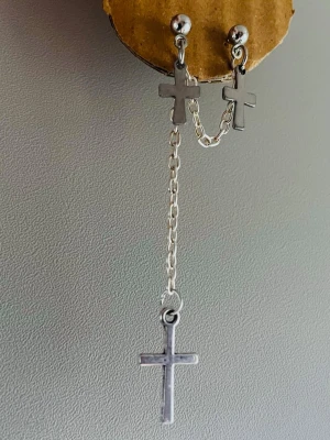 By prisförslag- 1 st Silverfärgat korsörhänge med kedja - Snyggt örhänge i silverfärgad metall med korsformade hängen och kedjedetaljer. Ena örhänget har en längre kedja med ett extra kors, vilket ger en cool och edgy look. Perfekt för dig som gillar statement-accessoarer och vill sticka ut.