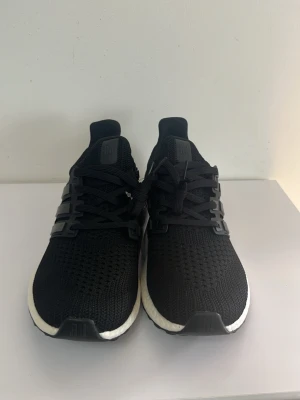 Svarta Adidas Ultraboost sneakers - Snygga svarta Adidas Ultraboost sneakers med stickad ovandel och klassiska tre ränder på sidan. Skorna har en vit, dämpad Boost-sula och svart yttersula. Perfekta för dig som gillar sportig och stilren look. Skön passform och modern design.