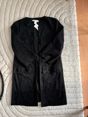 Svart lång kappa med fickor - Stilren svart kappa i mjukt material med raka linjer och två stora framfickor. Kappan har öppen front utan knappar och långa ärmar. Perfekt för dig som gillar minimalistisk och modern look.