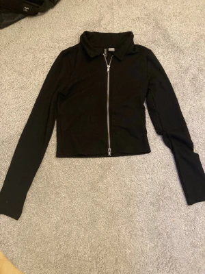 Svart kofta med dragkedja  - Fin svart kofta i ribbat material med silvrig dragkedja. Passar bra att styla med linnen! Använd men ännu i fint skick. 