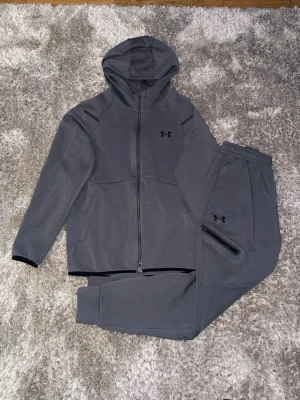 Under Armour Unstoppable  - Säljer ett grått träningsset från Under Armour med zip hoodie och matchande byxor. Setet har minimalistisk design, dragkedja framtill, huva och diskreta loggor på både jacka och byxa. Perfekt för gymmet eller chill. Materialet är mjukt och stretchigt för maximal komfort.