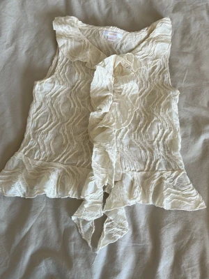 Vit ärmlös volangblus - Snygg vit ärmlös blus med volangdetaljer framtill och längs nederkanten. Blusen har ett vågigt, strukturerat tyg som ger en cool och unik look. Perfekt att styla med jeans eller kjol för en trendig outfit. Helt slutsåld. Skriv för fler bilder💘