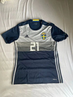 Svensk landslagströja Adidas #21 Durmaz  - Säljer en blå och grå svensk landslagströja från Adidas med nummer 21 och namn Durmaz på ryggen. Tröjan har korta ärmar, broderad svensk flagga på bröstet och klassiska Adidas-ränder på sidorna. Materialet är lätt och andas, perfekt för fotboll.