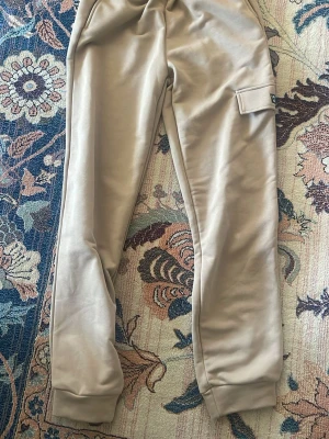 Beige mjukisbyxor från SHEIN - Snygga beige mjukisbyxor från SHEIN med elastisk midja och muddar vid benslut. Byxorna har en praktisk ficka på sidan och är tillverkade i mjukt material som är skönt att ha på sig. Perfekta för chill dagar och avslappnade outfits.