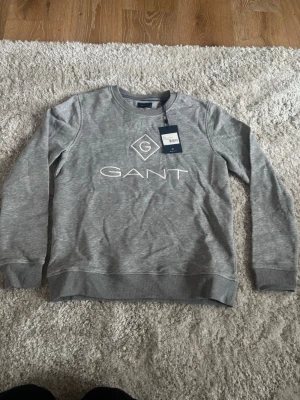 Grå sweatshirt från GANT - Snygg grå sweatshirt från GANT med klassisk logga tryckt framtill. Tröjan har rund halsringning, ribbade muddar vid ärmslut och nederkant samt en avslappnad passform. Skulle säga att den är rätt liten i storleken