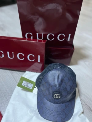 Mörkblå Gucci keps med GG-mönster - Snygg mörkblå keps från Gucci med klassiskt GG-monogram över hela kepsen och metallogo framtill. Justerbar rem bak och mesh-paneler på sidorna för extra ventilation. Perfekt accessoar för dig som vill ha en exklusiv och trendig look.