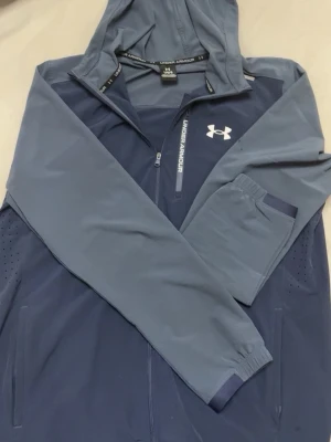 Blå hoodie från Under Armour - Snygg tvåfärgad Jacka från Under Armour i mörkblått och ljusblått. Dragkedja framtill, huva och fickor på sidorna. Tillverkad i ett lätt och sportigt material, perfekt för en aktiv stil. Logga på bröstet och diskreta detaljer längs ärmarna.