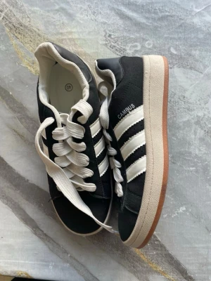 Adidas Campus svarta sneakers - Klassiska Adidas Campus sneakers i svart mocka med vita läderdetaljer och de ikoniska tre ränderna på sidan. Tjock vit sula med brun yttersula och vita skosnören. CAMPUS-tryck i vitt på sidan. Perfekta för en clean och stilren look.