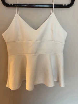 Vit peplum linnetopp med smala band - Snygg vit linnetopp med smala axelband och v-ringning. Toppen har en markerad midja och peplumformad nederdel som ger en feminin siluett. Perfekt att styla med jeans eller kjol för en trendig look.