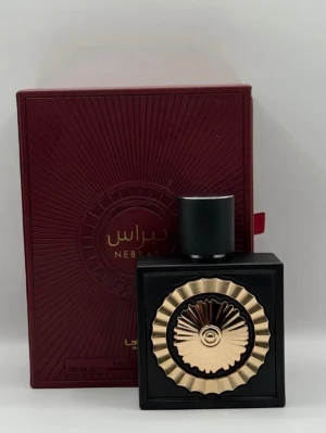 Lattafa Nebras Eau de Parfum 100ml - Lattafa Nebras Eau de Parfum kommer i en elegant svart fyrkantig flaska med en unik guldig dekor på framsidan och svart lock. Förpackningen är mörkröd med gulddetaljer och arabiska bokstäver. Flaskan rymmer 100 ml och har en lyxig och modern design som sticker ut i parfymhyllan.