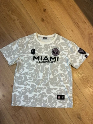  BAPE x Inter Miami T-shirt  - Säljer en limited edition t-shirt från A Bathing Ape i samarbete med Inter Miami. T-shirten har ett vitt och grått camo-mönster, tryck med MIAMI och BAPE-logga på bröstet samt patch på ärmen. Perfekt för dig som gillar streetwear och fotboll.