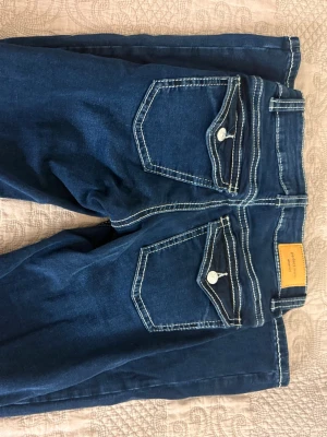 Mörkblå bootcut jeans med kontrastsömmar - Säljer ett par mörkblå bootcut jeans från Gina tricot med tydliga vita kontrastsömmar och snygga fickdetaljer bak. Jeansen har låg midja och klassisk femficksmodell. Materialet är denim med en mjuk känsla och stretch för extra komfort.