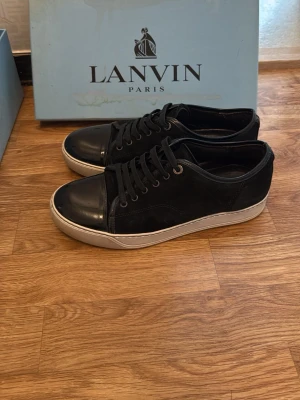 Svarta Lanvin sneakers med vit sula - Eleganta Lanvin Cap Toe i svart lackläder med klassisk och tidlös design. Den blanka finishen och cap toe-detaljen gör dem perfekta för formella tillfällen eller en dressad stil. Kvitto, 2 påsar, låda och extra snören finns med. Pris går att diskutera!  	•	Storlek: UK 6 	•	Skick: 7/10 (mycket bra skick med mindre tecken på användning) 	•	Färg: Svart