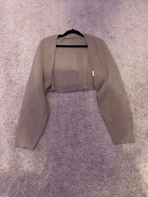 Beige ribbad bolero med långa ärmar - Kort beige bolero i ribbstickat material med långa ärmar och öppen front. Perfekt att slänga över en topp för en chill och trendig look. Passar till både jeans och klänning för en avslappnad vibe.