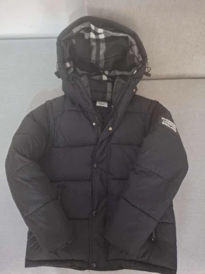 Svart pufferjacka Burberry England - Snygg svart pufferjacka från Burberry England med stor huva och rutigt foder. Jackan har dragkedja och knappar framtill samt logga på ärmen. Perfekt för kalla vinterdagar och riktigt trendig look.