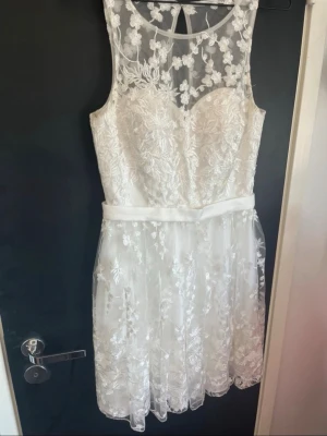 Vit spetsklänning med blommönster - Kort vit klänning med vackra spetsdetaljer och blommigt mönster. Klänningen har ärmlös design, markerad midja med ett brett band och transparent överdel med broderade blommor. Perfekt för sommarens speciella tillfällen.