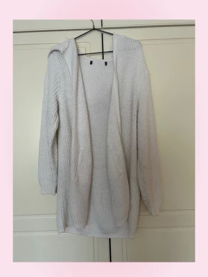 Vit stickad hoodiekofta - Mysig vit stickad kofta med huva och långa ärmar. Koftan har öppen framsida och ribbstickad struktur, perfekt för lager på lager. Passar dig som gillar en avslappnad och enkel stil. ( nopprig )