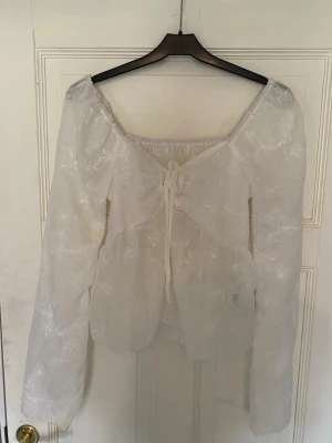 Vit offshoulder blus med spetsdetaljer - Romantisk vit offshoulder blus med långa ärmar och fina broderade spetsdetaljer. Blusen har snörning framtill och smockad rygg för en bekväm passform. Tunn och luftig, perfekt för en trendig och feminin look.