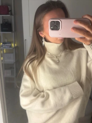 Beige stickad tröja med hög krage - Säljer en beige stickad tröja med hög ribbad krage och långa ärmar. Tröjan har bred ribbstickad mudd vid nederkant och ärmar, samt en avslappnad passform. Perfekt för kalla dagar och enkel att styla till olika outfits.