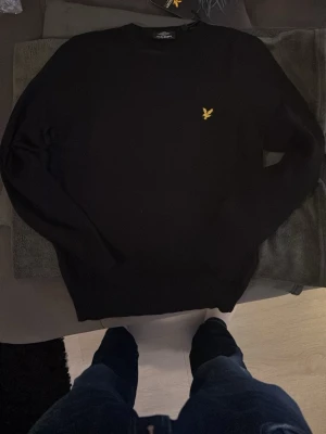 Svart stickad tröja från Lyle & Scott - Klassisk svart stickad tröja från Lyle & Scott med rund hals och den ikoniska gula örnloggan broderad på bröstet. Tröjan har en enkel och stilren design som passar till det mesta. Perfekt basplagg för garderoben.