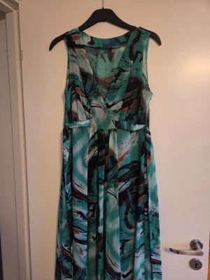 Mönstrad långklänning i grönt och blått - Säljer en långklänning med abstrakt mönster i gröna, blå och bruna toner. Klänningen har v-ringning, är ärmlös och har ett omlottliv. Perfekt för dig som vill sticka ut med en färgstark look.