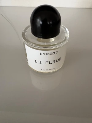 Byredo Lil Fleur Eau de Parfum - Byredo Lil Fleur Eau de Parfum i en rund glasflaska med minimalistisk vit etikett och svart rund kork. Doften är elegant och flaskan har en stilren design som passar perfekt på parfymhyllan. Materialet är glas och plast. Ca 20/25 ml kvar 