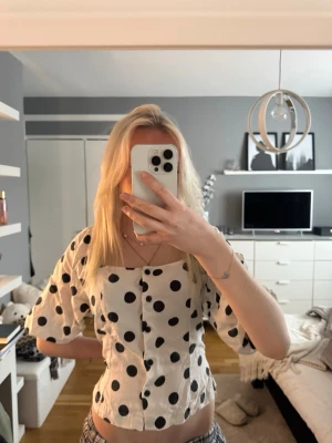 Vit prickig blus med puffärm - Supersöt vit blus med stora svarta prickar och puffiga trekvartsärmar. Blusen har en lätt croppad passform och knappar framtill. Perfekt för dig som vill sticka ut lite extra med ett lekfullt mönster. 🖤🤍