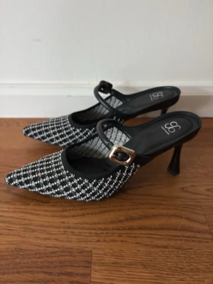 Svartvita pumps med rutmönster - Nya oanvända syäuper snygga sköna skor. Små i strl us8 är som strl 38. Säljer ett par snygga pumps från Cosy Island  med svartvitt rutmönster och spetsig tå. Skorna har en elegant rem med silverfärgat spänne och smal klack. Materialet är textil med mjuk insida och klassisk design som sticker ut.