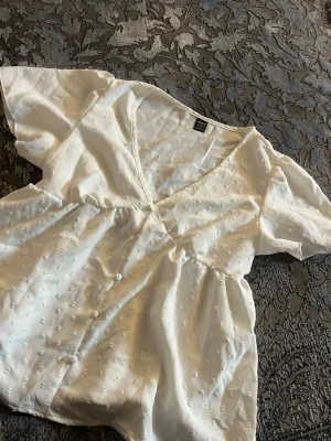 Vit blus med knappar  - Söt vit blus med korta puffärmar och v-ringning. Blusen har dekorativa knappar framtill och ett diskret prickigt strukturmönster i tyget. Tillverkad i ett lätt och luftigt material som ger en avslappnad känsla storlek S och aldrig använts 