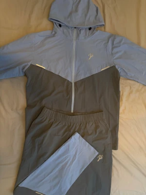 Gråblå tracksuit från jeu d'ombres  - Snygg tracksuit från  jeu d'ombres i tvådelad design med jacka och byxor. Färgkombination i ljusblått och mörkgrått, jackan har huva och dragkedja samt två fickor med dragkedja. Byxorna har resår i midjan och matchande färgblock. Perfekt för träning eller chill.