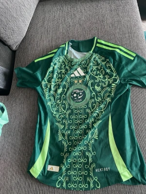 Grön Adidas fotbollströja Saudiarabien - Snygg grön fotbollströja från Adidas med Saudiarabiens landslagsemblem och dekorativa mönster i ljusgrönt på framsidan. Tröjan har korta ärmar, rund hals och klassiska Adidas-ränder på axlarna. Tillverkad i lätt och ventilerande funktionsmaterial.