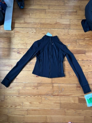 Svart träningsjacka från Lululemon - Säljer en svart träningsjacka från Lululemon med figurnära passform och långa ärmar. Jackan har dragkedja framtill, två fickor med dragkedja och snygga sömdetaljer. Perfekt för gymmet eller löpning. Materialet är stretchigt och funktionellt.