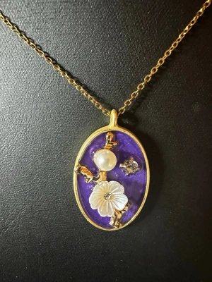 Lila halsband med pärldetaljer - Unikt halsband med en oval, guldfärgad ram och lila bakgrund. Hänget har en vit pärla, en snidad blomma och små gulddetaljer. Kedjan är tunn och guldfärgad. Perfekt för dig som gillar statement-smycken med vintagekänsla.