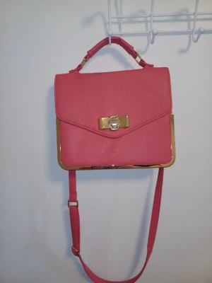 Rosa axelväska med gulddetaljer - Snygg rosa axelväska med guldfärgade metalldetaljer och vridlås framtill. Väskan har både ett justerbart axelband och ett handtag upptill. Stilren och boxig form som ger en trendig look. På andra bilden ser man att den har en vit fläk där bak o på tredje bilden så ser man att handtaget är lite sisådär, skriv om ni har frågor, priset går att diskutera ❤️