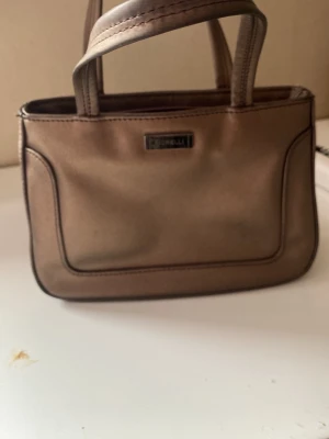 Brun handväska från Fiorelli - Snygg brun handväska från Fiorelli med stilren design och två handtag. Väskan har en metallbricka med loggan på framsidan och är gjord i ett glansigt material som ger en lyxig känsla. Perfekt storlek för det viktigaste och enkel att matcha till olika outfits.